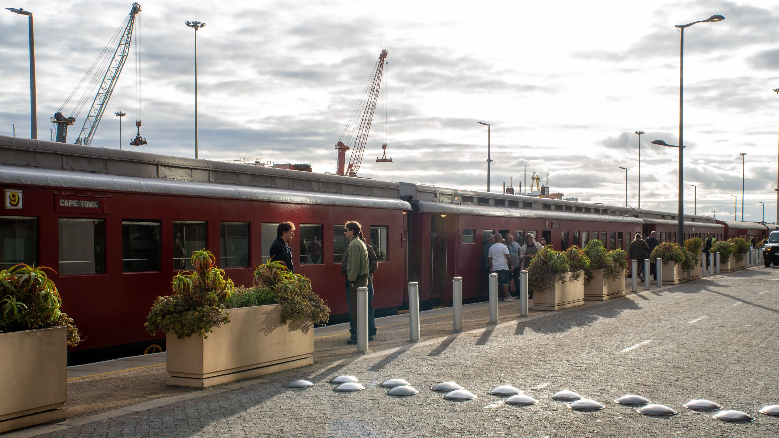 People wait beside a red train at a platform with large planters.