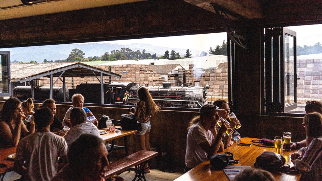 People seated indoors at tables, drinking; view of a vintage train outside the window.