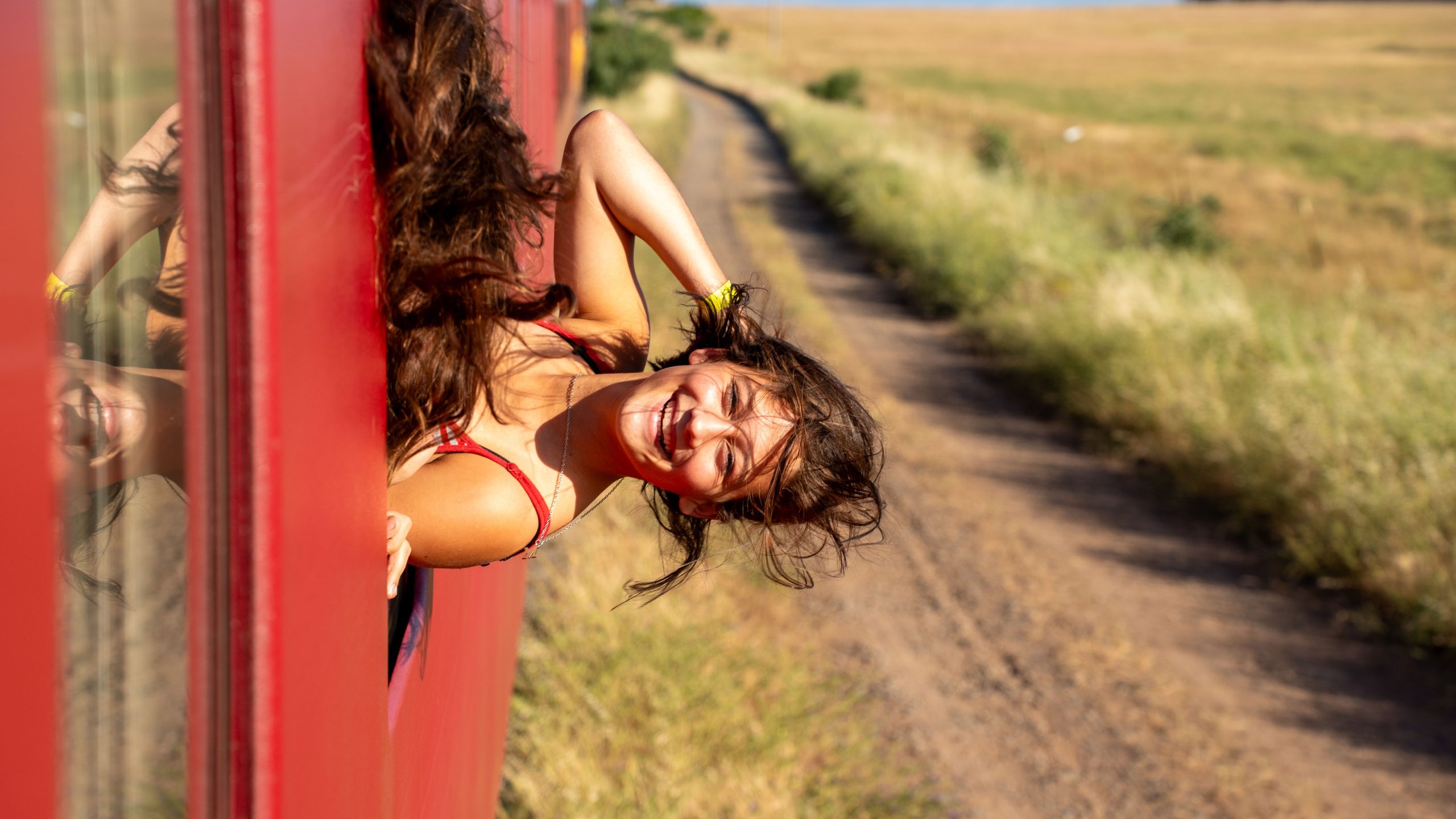 Smiling person leaning out of a red train on a countryside track.