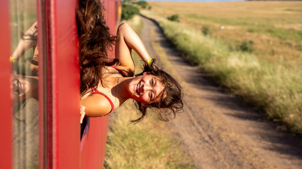 Smiling person leaning out of a red train on a countryside track.