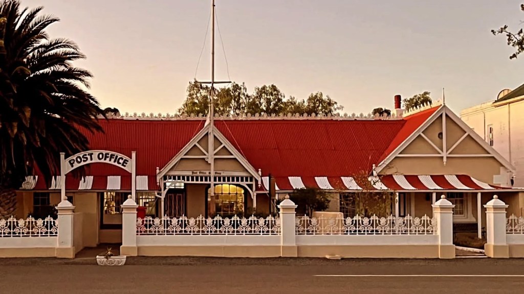 Vintage post office with red roof and striped awnings, surrounded by a white fence.
