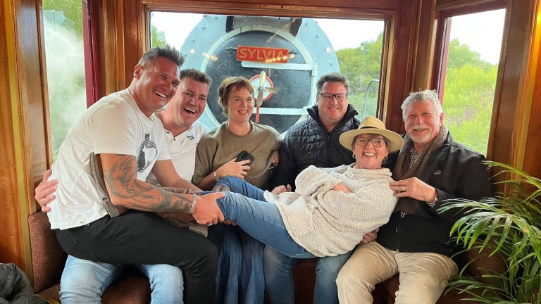Six people smiling on a train with a steam engine background.