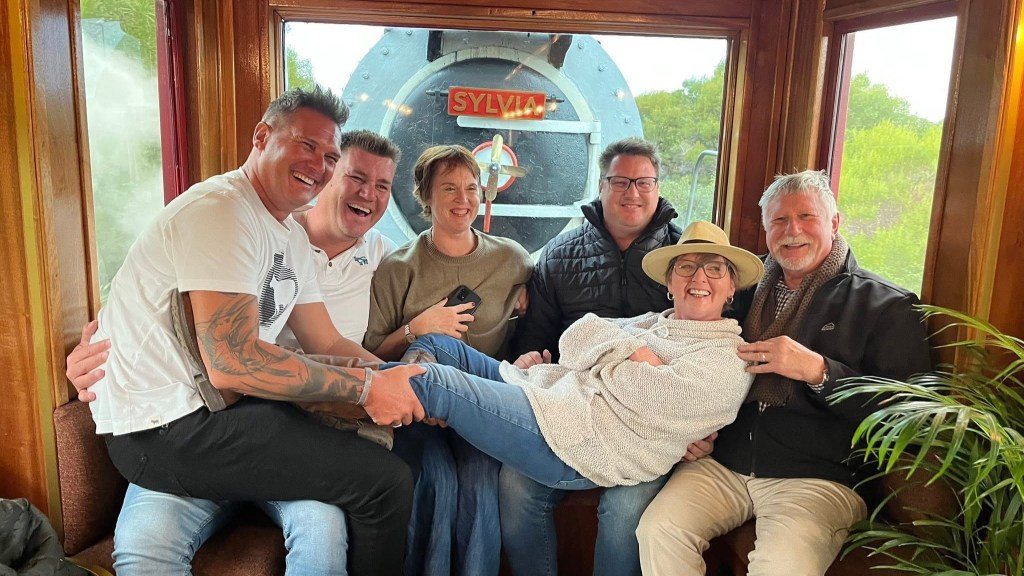 Six people smiling on a train with a steam engine background.