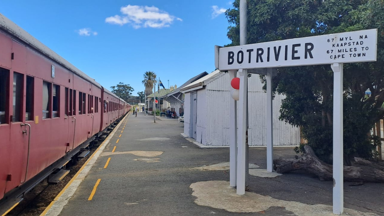 Red train at Botrivier station with sign to Cape Town.