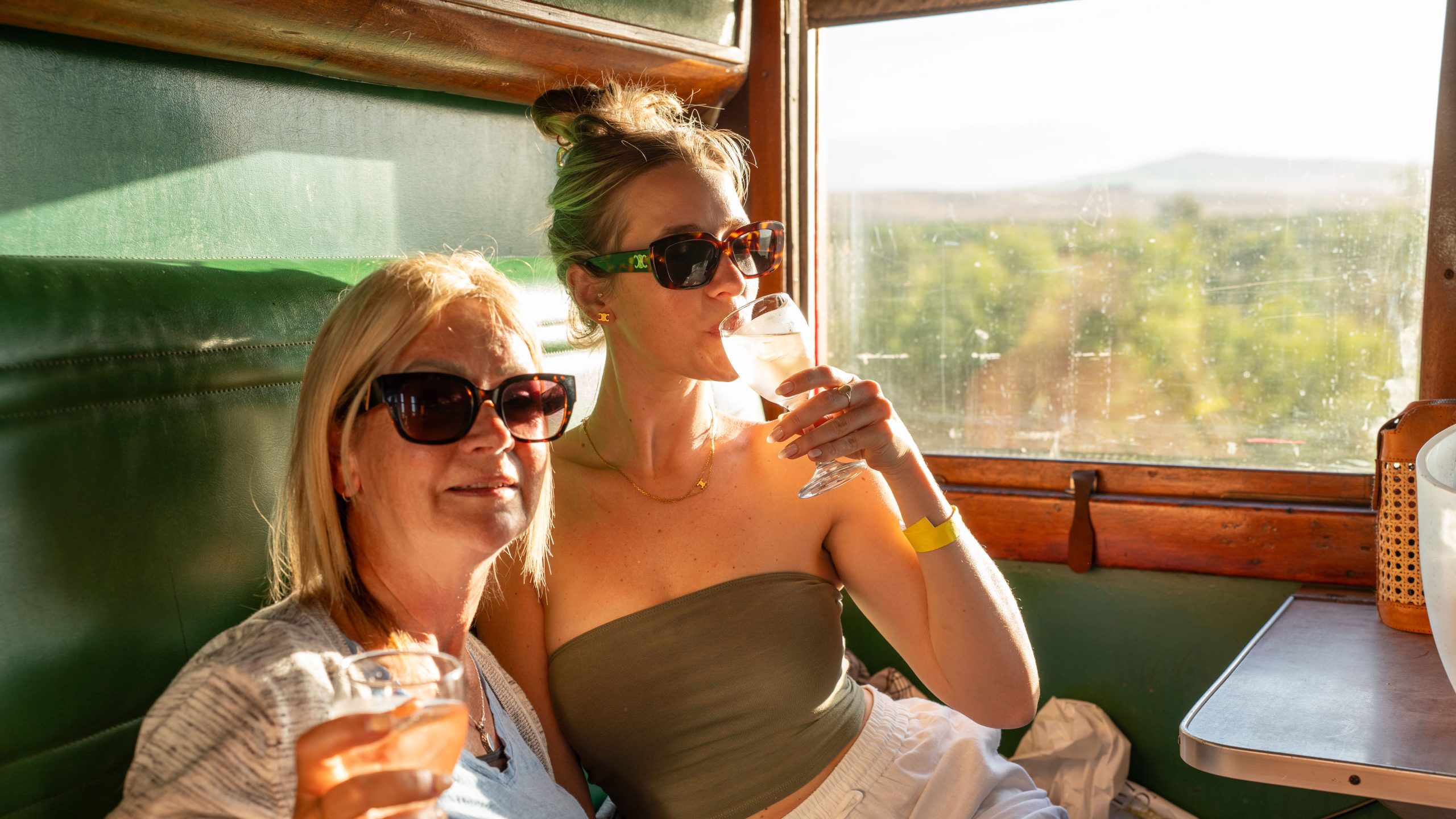 Two women in sunglasses enjoying drinks on a train with a scenic view outside the window.