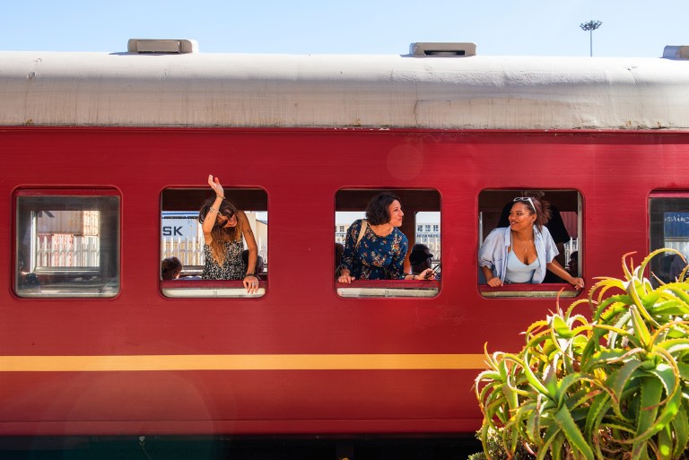Three people waving from windows of a red train at a station, plants in the foreground.