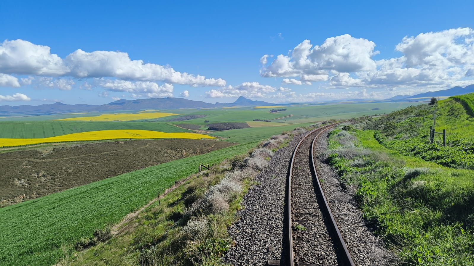 Curved railway track through rolling green and yellow fields under a blue sky with clouds.