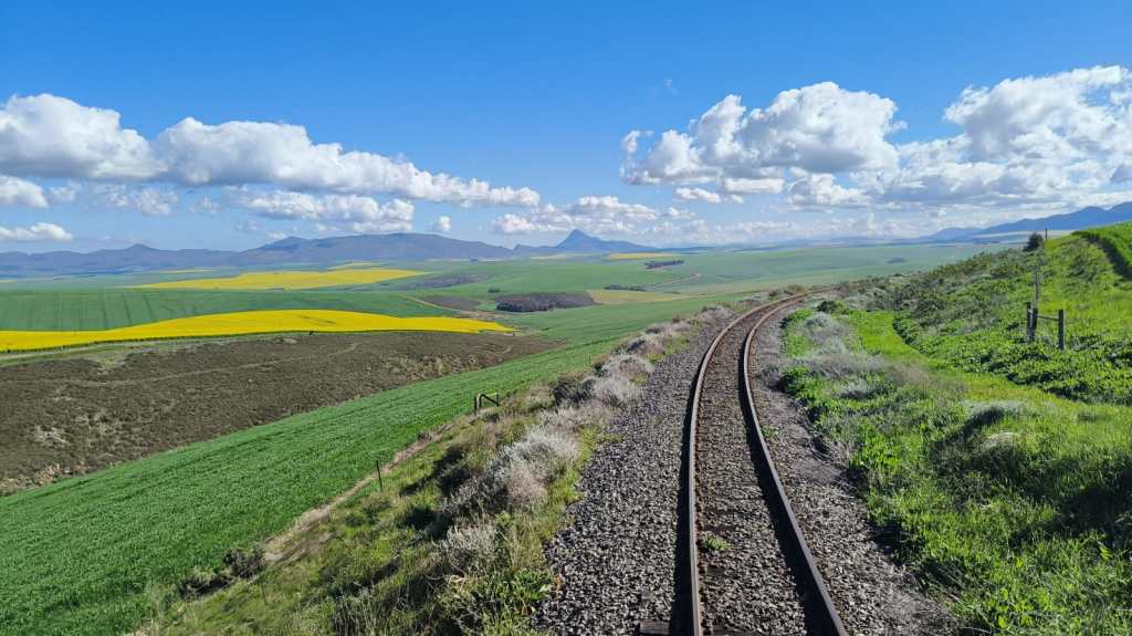 Curved railway track through rolling green and yellow fields under a blue sky with clouds.