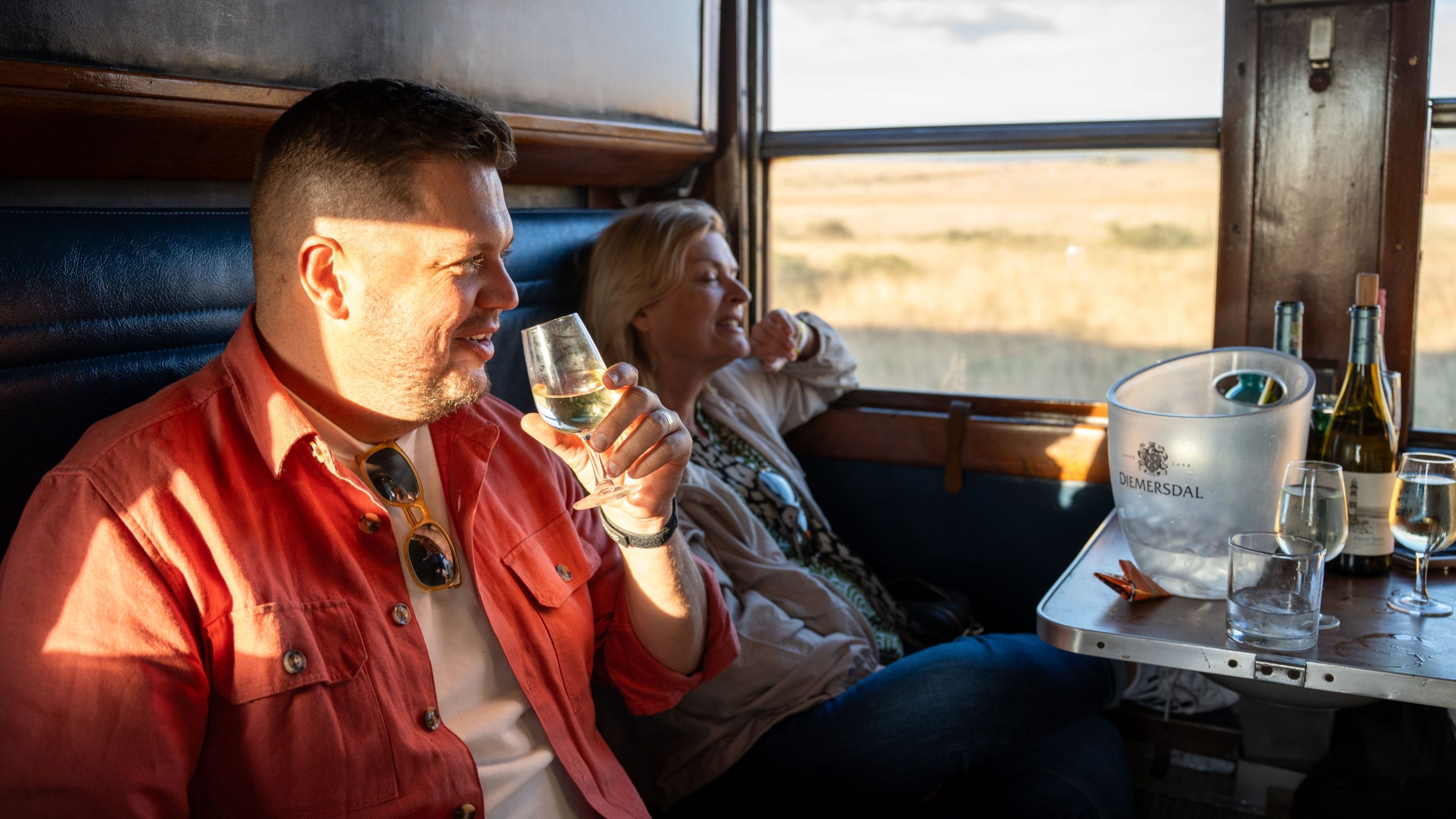 Two people enjoy drinks in a train cabin with a window view.