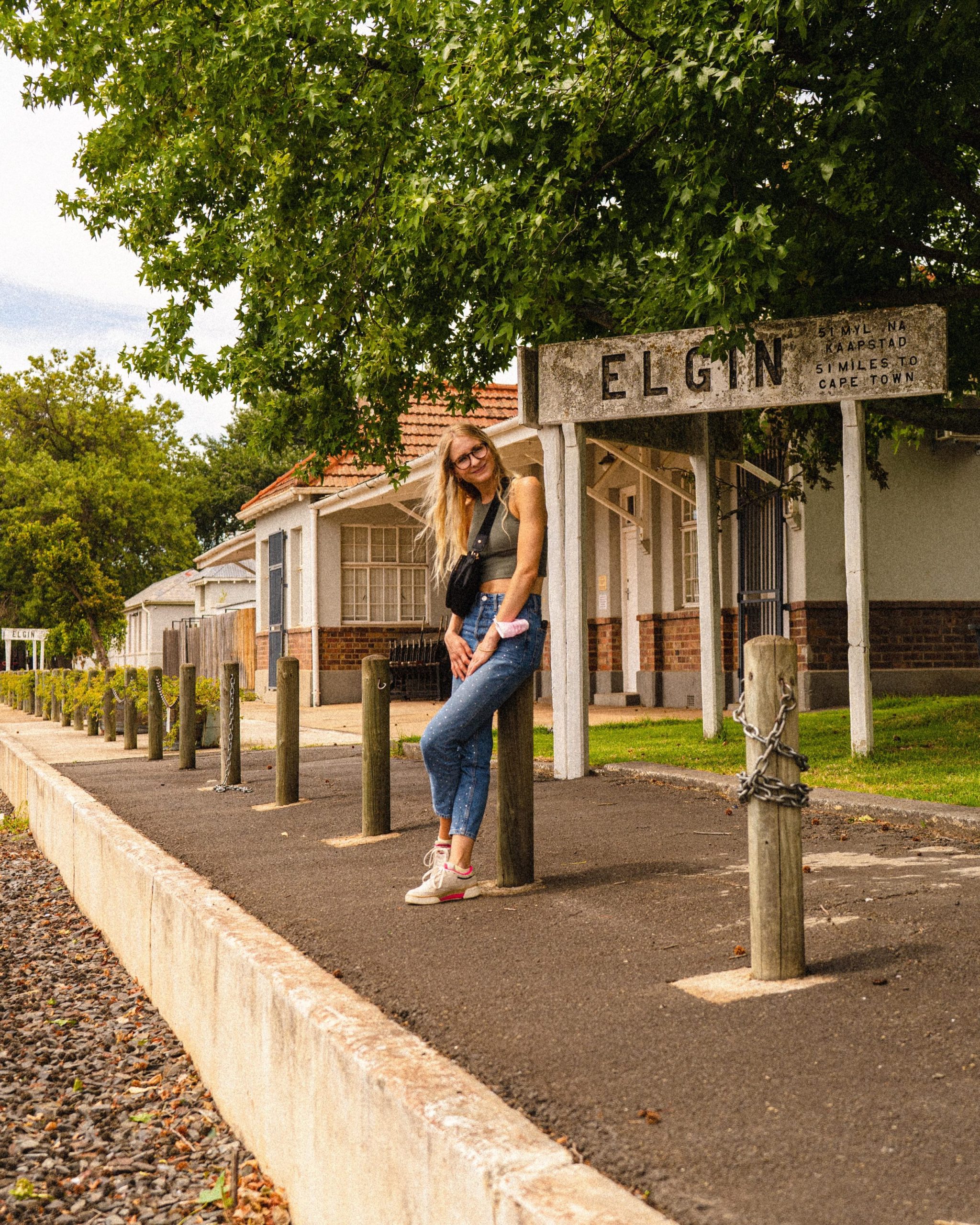 Person posing beside Elgin train station sign, trees and buildings in background.