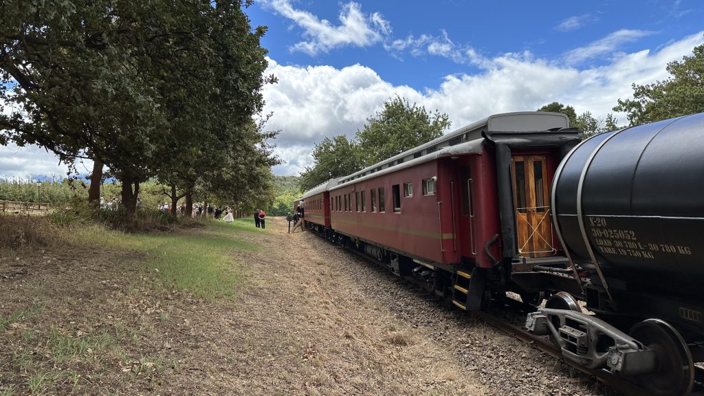 Red train on tracks under a blue sky with clouds and trees nearby.