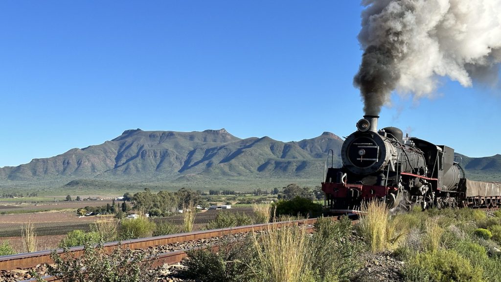 Steam train on tracks with mountains in the background under a clear blue sky.