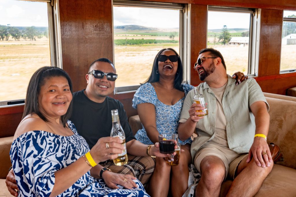 Four people sitting on a train, smiling and holding drinks.