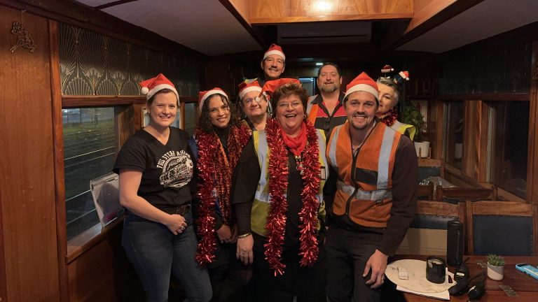 Group of people in festive attire with Santa hats and tinsel, posing inside a wooden room.