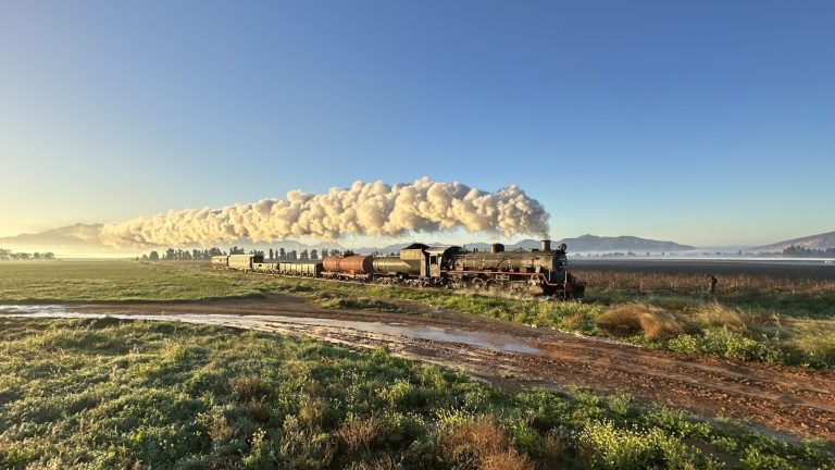 Steam train with long smoke trail crosses scenic landscape under clear blue sky.