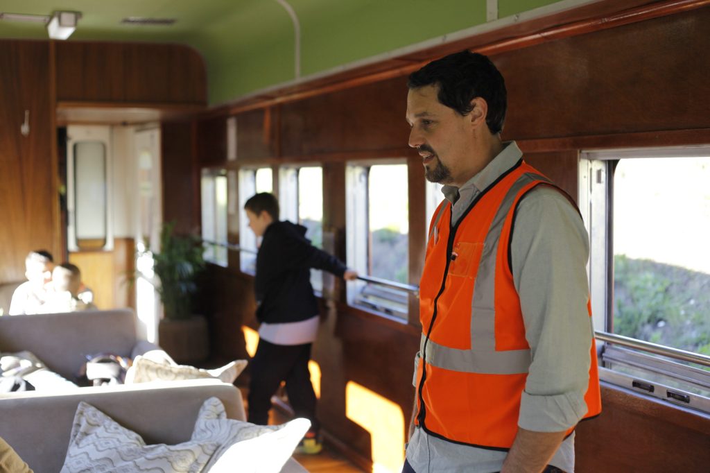Man in orange vest in train car with wood paneling, passengers seated.
