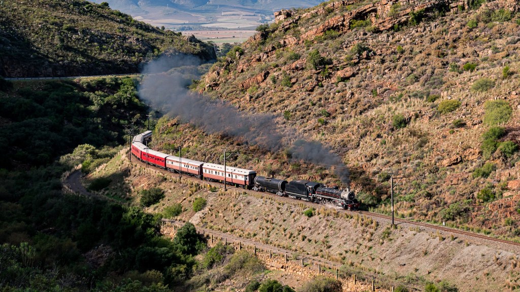 Steam train with red carriages traveling through a scenic mountain pass.