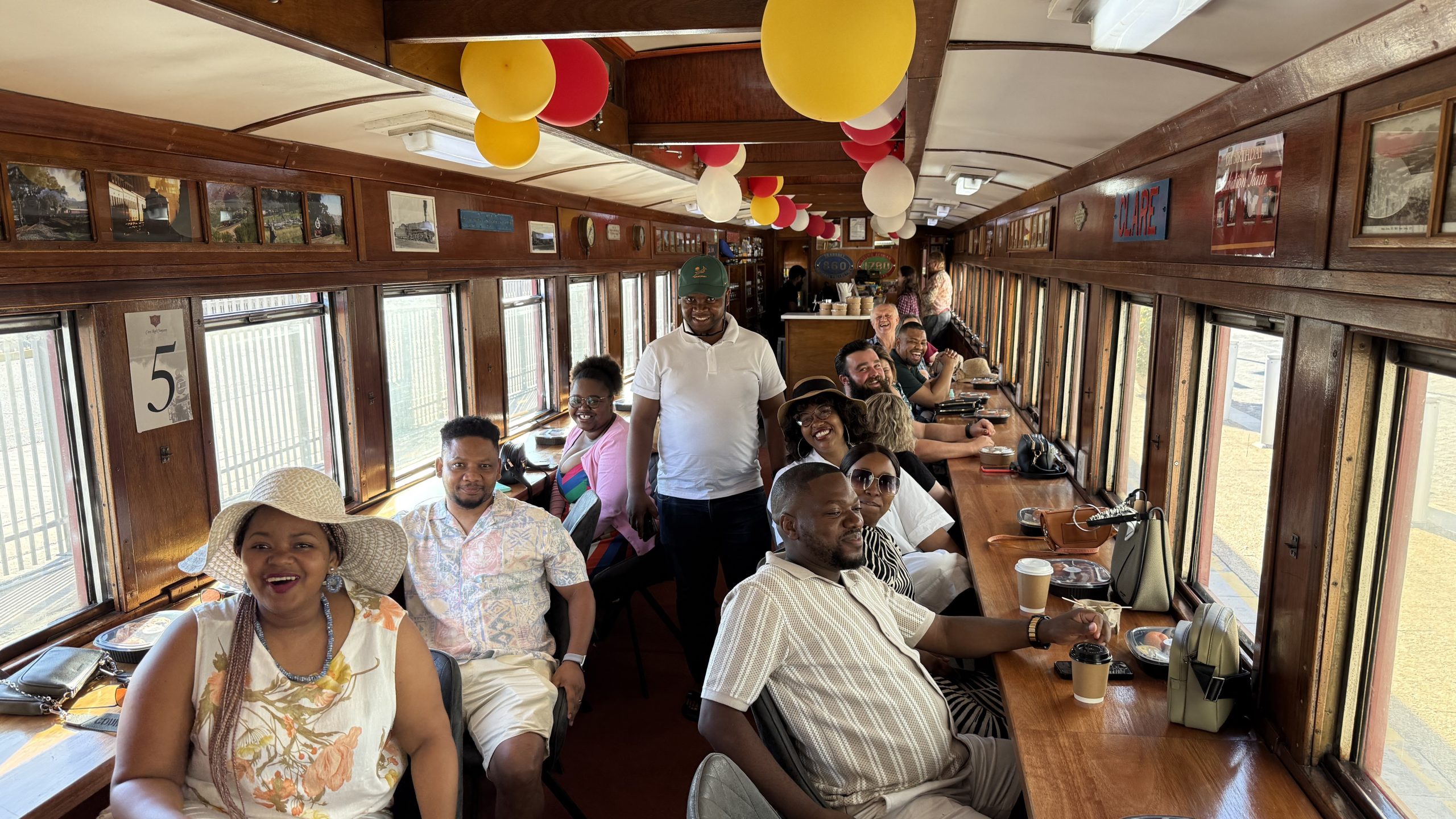 Group of people seated in a train car with balloons and sunlight through windows.