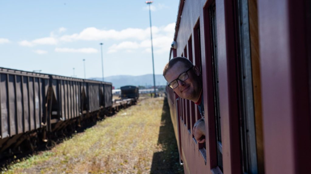 Person smiling, looking out train window, train tracks on left.