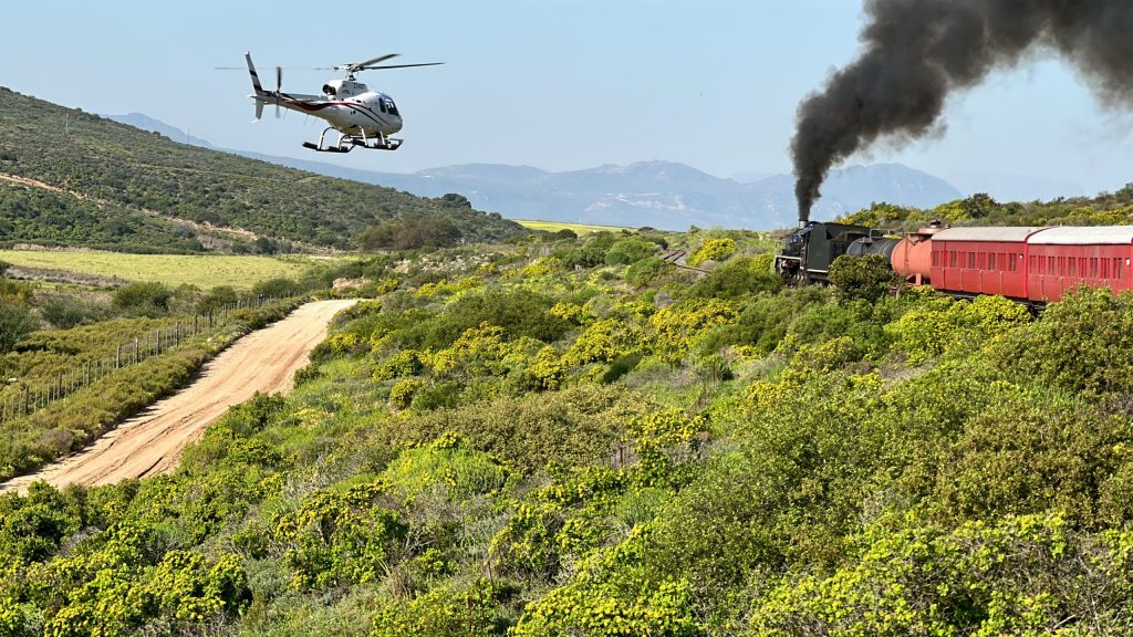 Helicopter flying near steam train with red carriages on a scenic hillside track.