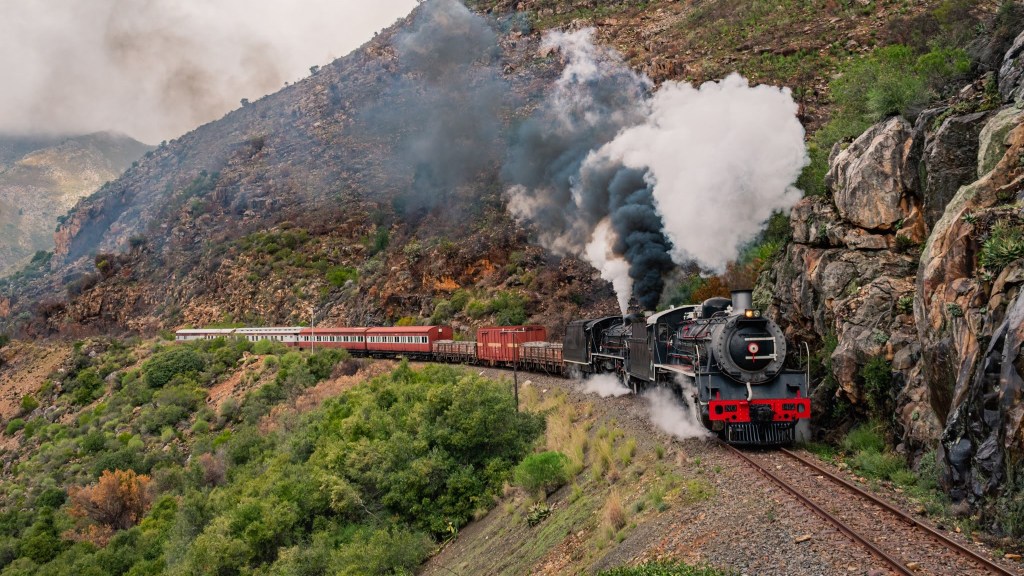 Steam train with smoke on a mountain track amid green and rocky scenery.