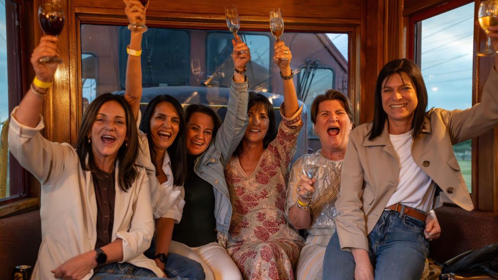 Six women smiling and raising wine glasses in celebration on a train.
