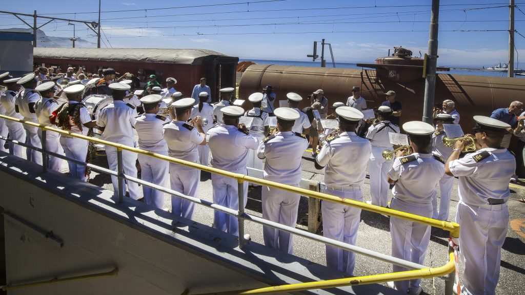 Navy band in white uniforms playing on a ship deck with trains in the background.