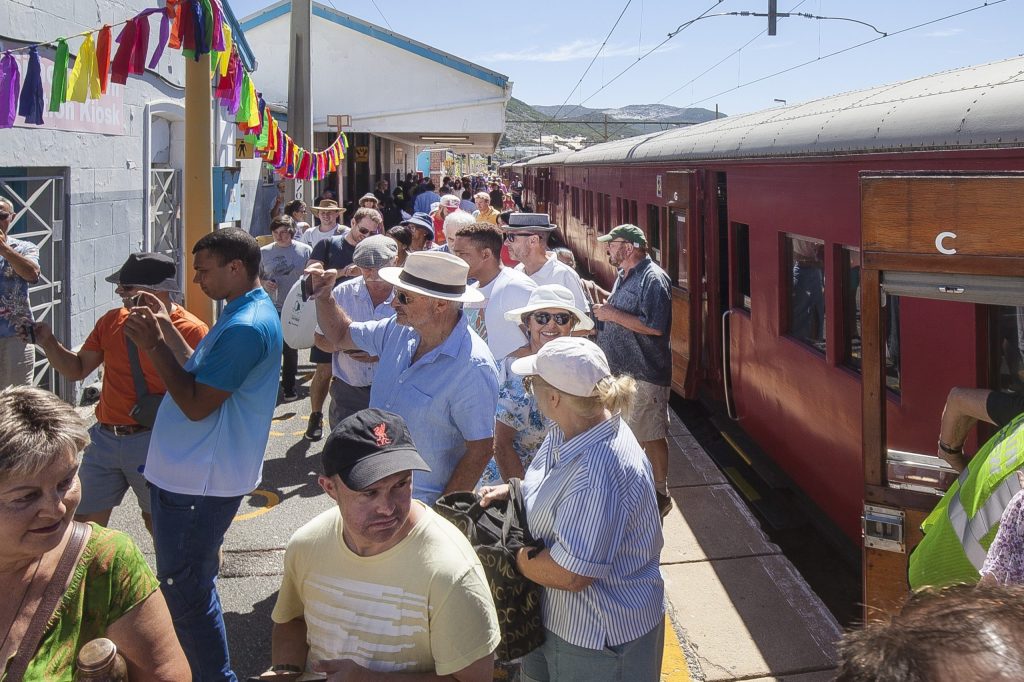 Crowd at train station near red train, colorful flags above.