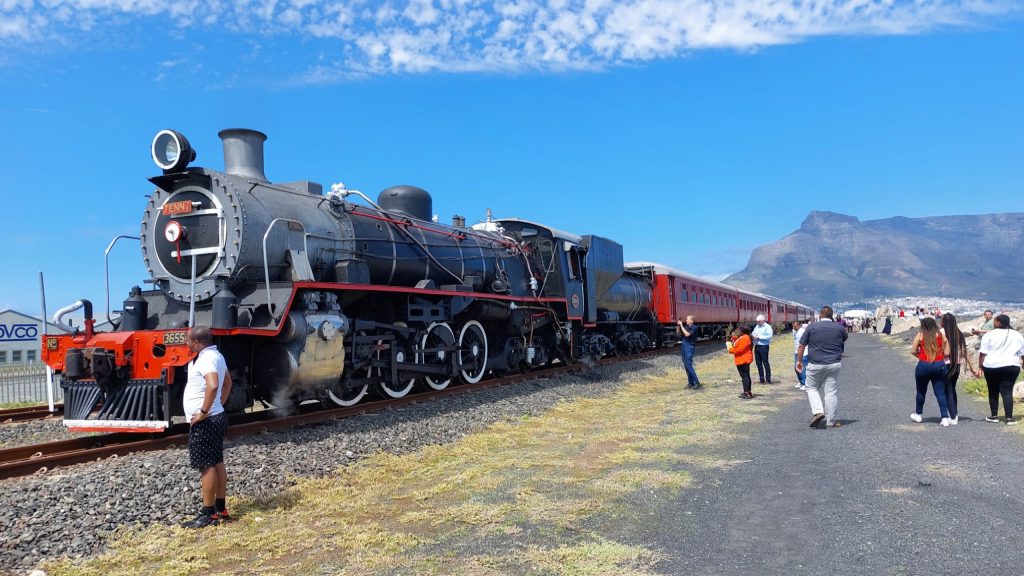 Vintage steam train on tracks with people taking photos, mountains in the background.