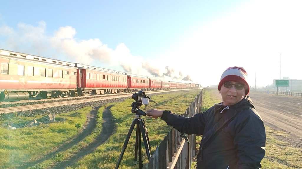 Person photographing a red train with a camera on a tripod beside a grassy field.