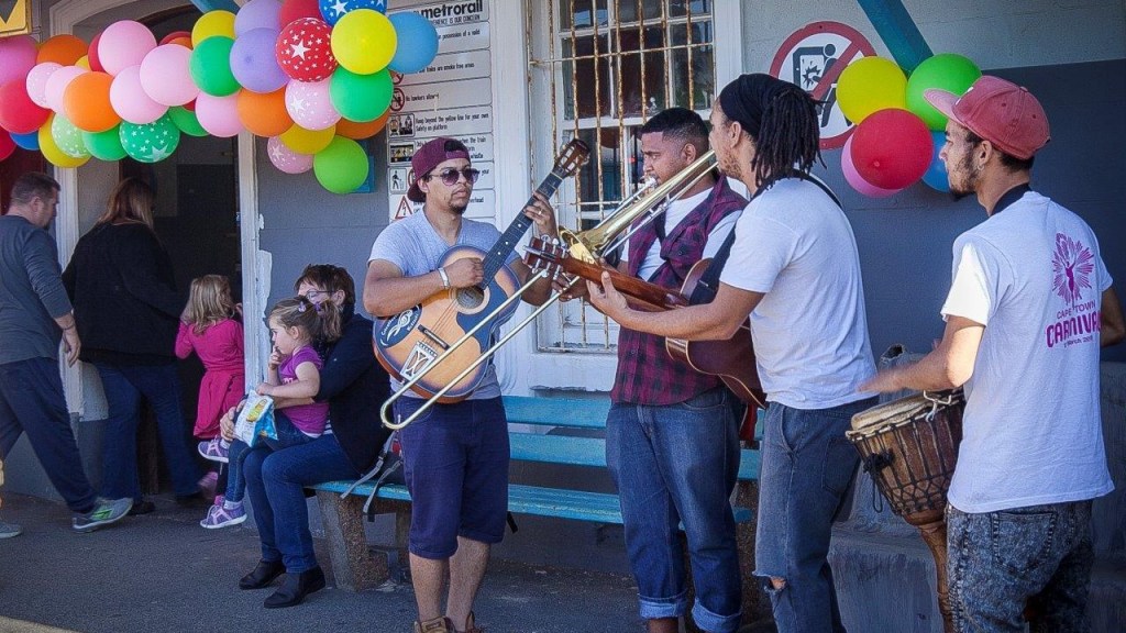 Street musicians play under balloons, people standing nearby on a sunny day.