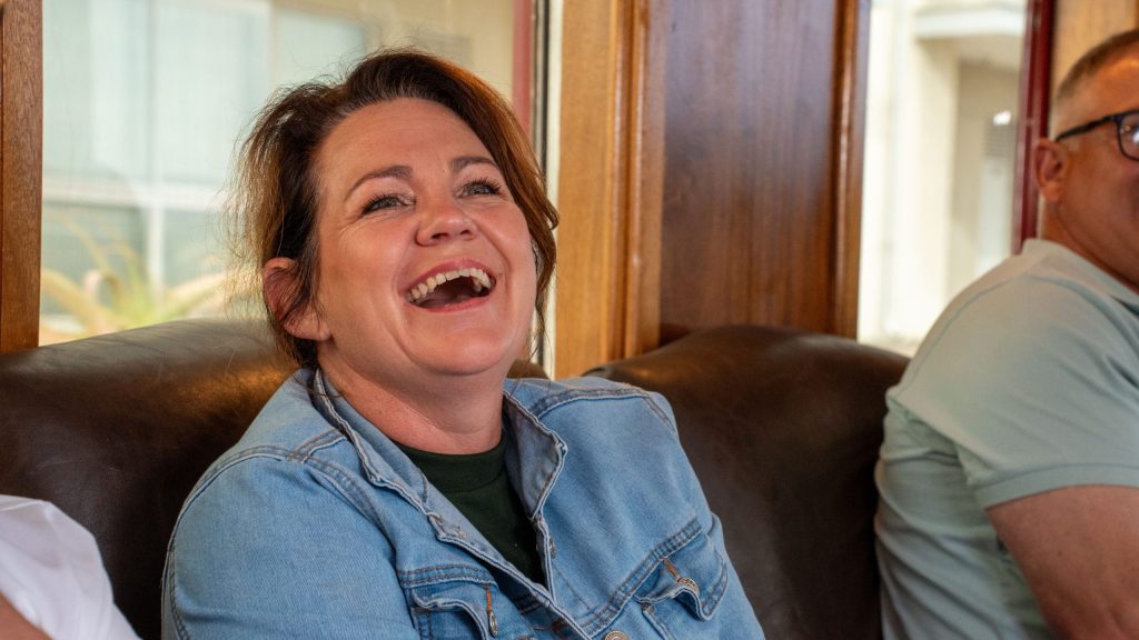 Woman in denim jacket laughing on a couch with others nearby.