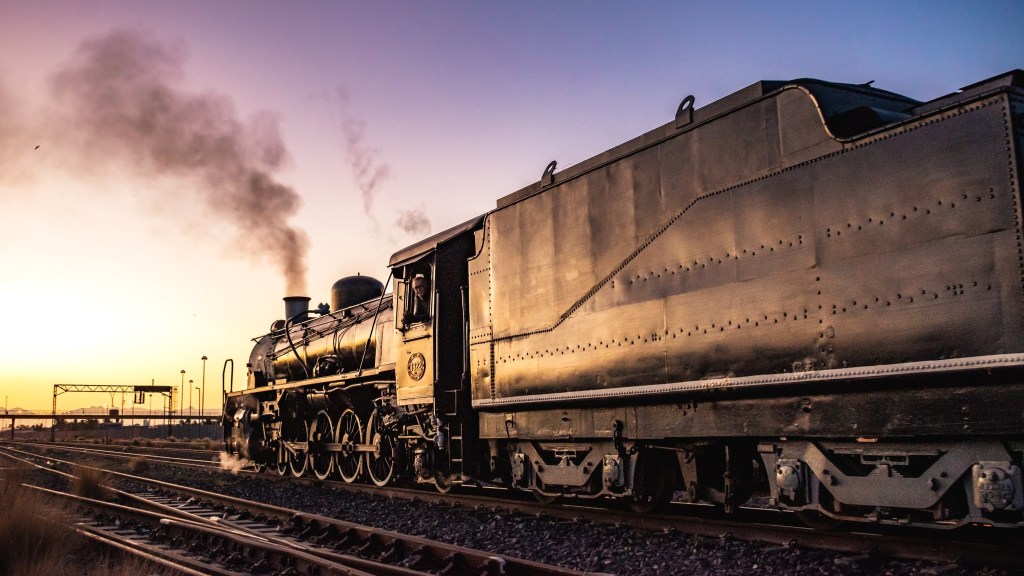 Steam train traveling on tracks at sunset with smoke billowing from chimney.