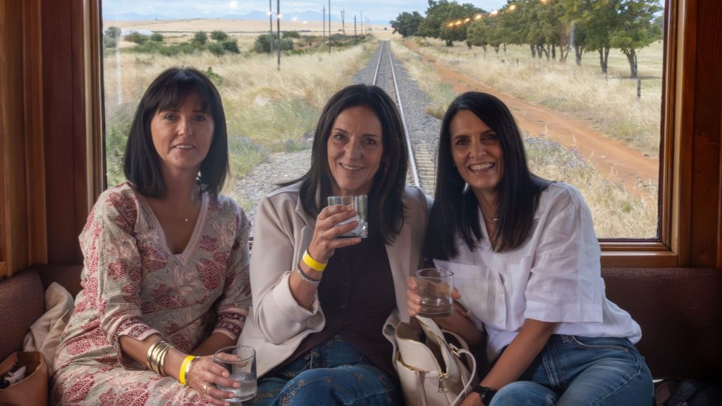 Three women sitting in a train with drinks, tracks visible behind.
