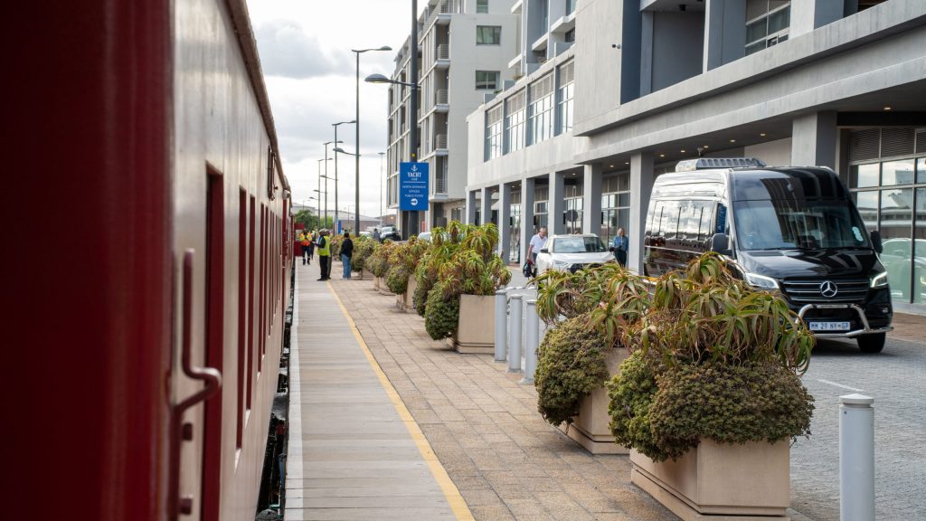 Train on left, sidewalk with plants and a black van on right, modern buildings in background.