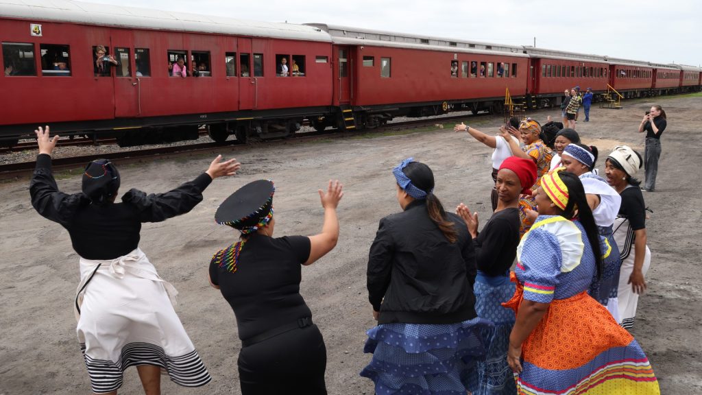 Group of people in colorful clothing waving at a red train with passengers.