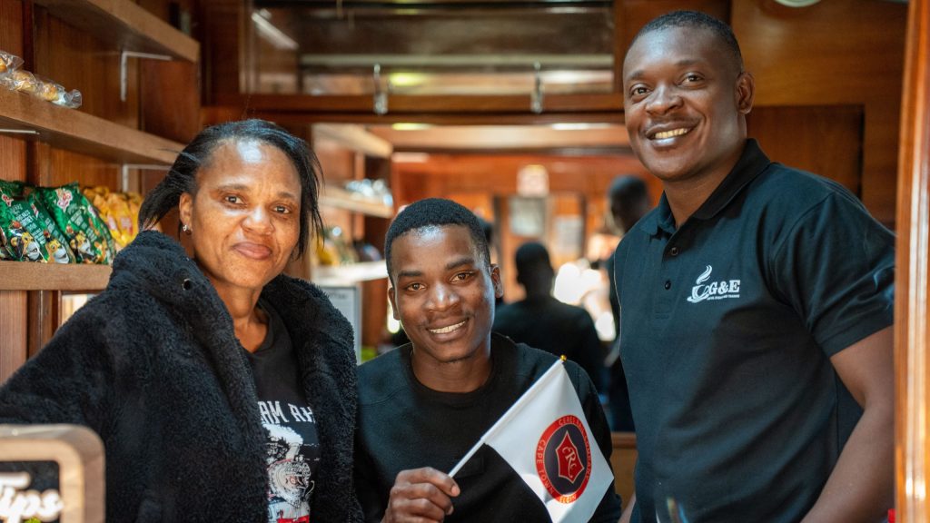 Three people smiling inside a shop, one holds a small flag with a logo.