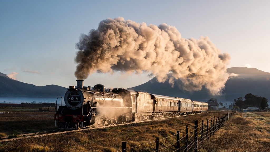 Steam train with billowing smoke on tracks in a rural landscape.