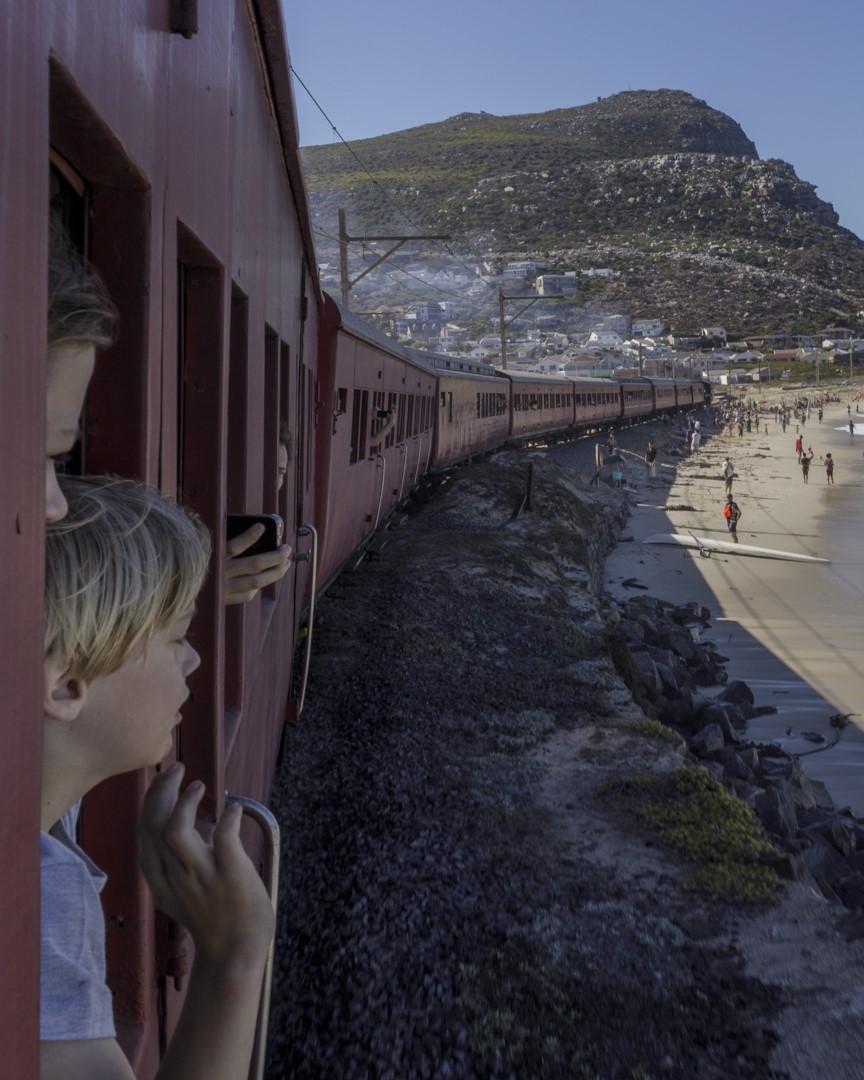 Train on coastal track with passengers looking out, beach and mountain in background.