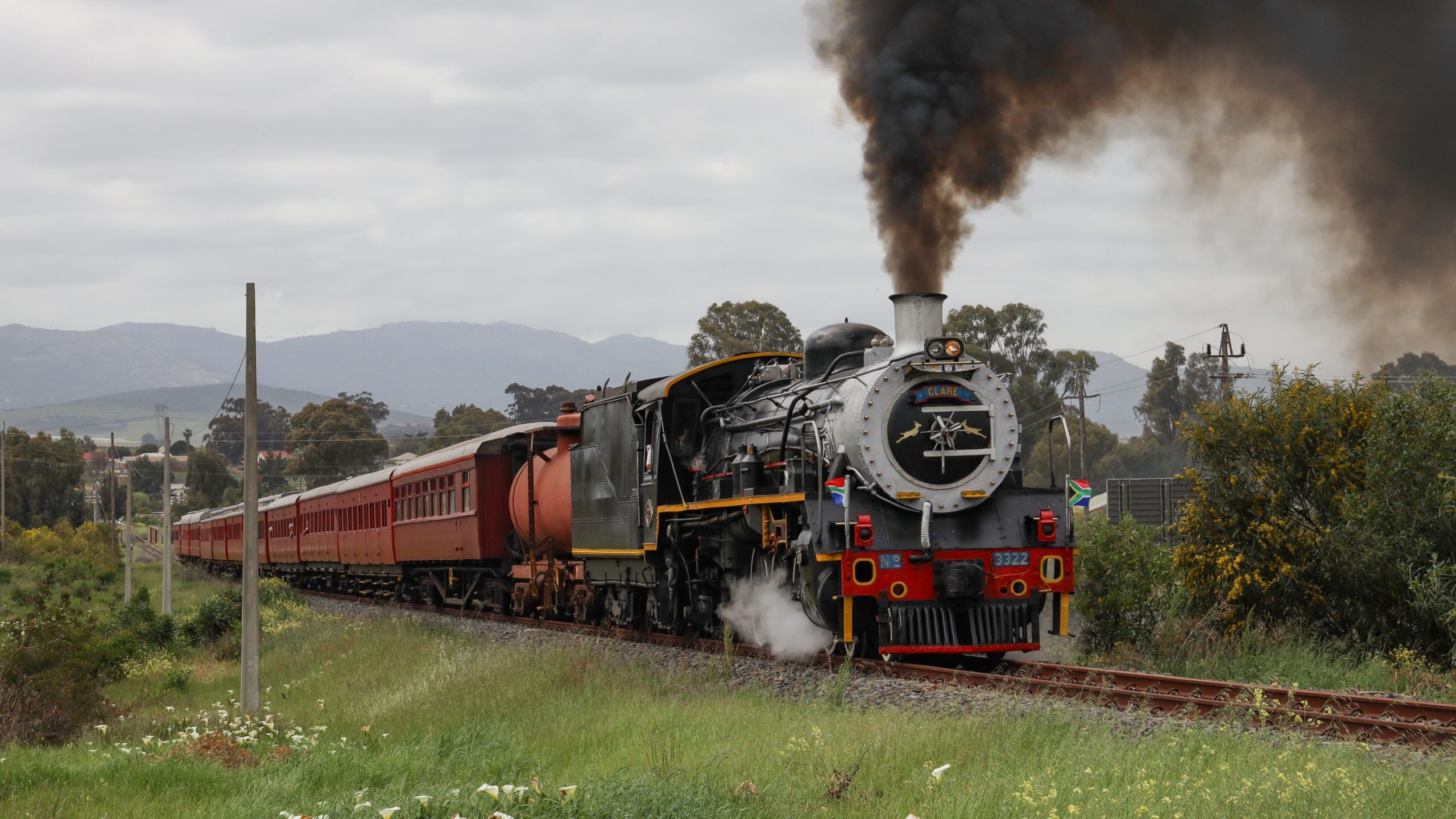 Steam train with red carriages on a rural track under cloudy sky.