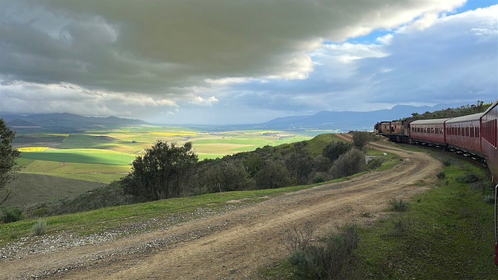 Red train on curved track with green fields and cloudy sky.