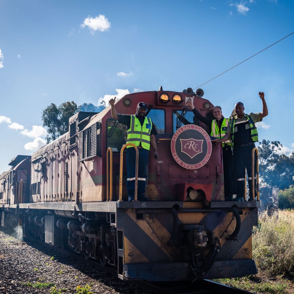 Locomotive with two people in safety vests standing at the front, one waving.
