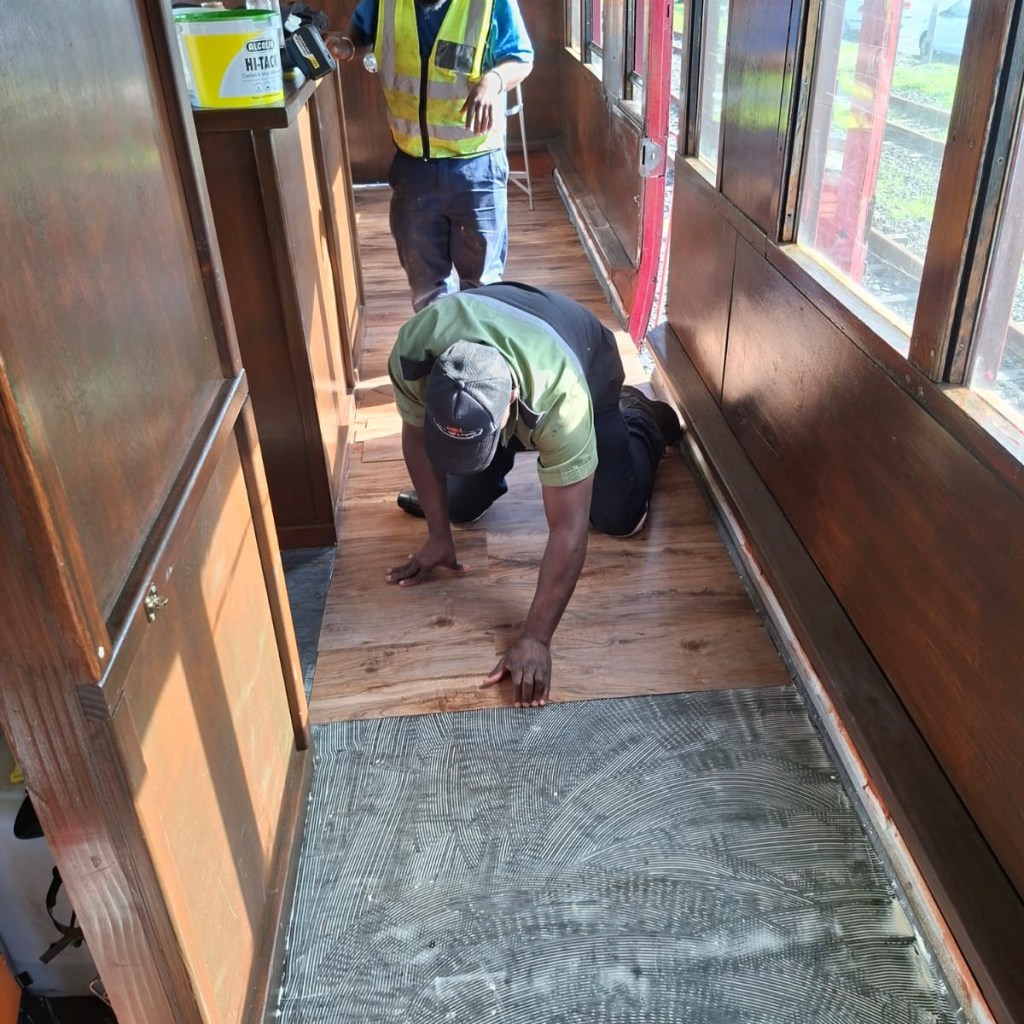 Two workers installing wood flooring in a narrow hall.